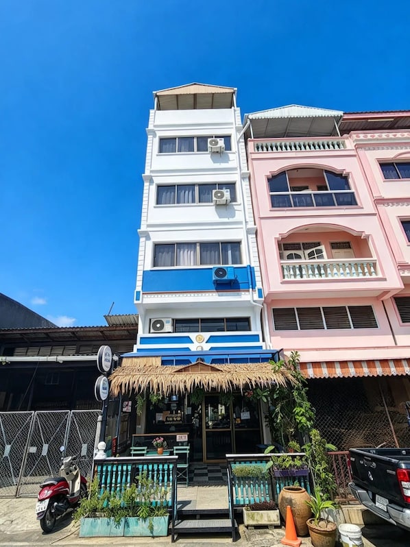 A multi-story building with a blue and white facade, a thatched-roof structure in front, and a parked motorcycle. - MT FRIEND HOME (Phuket)