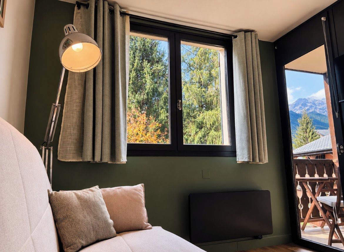 Un dormitorio con cama, lámpara de noche, ventana con cortinas y vistas a árboles y montañas.