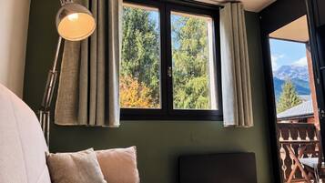 Un dormitorio con cama, lámpara de noche, ventana con cortinas y vistas a árboles y montañas.