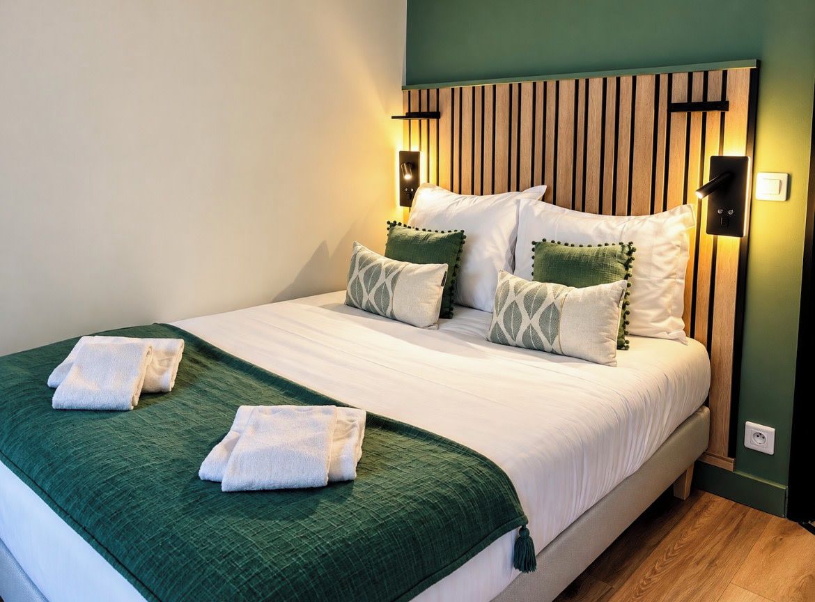 A neatly made bed with a green blanket and two towels, a wooden headboard with vertical slats, and two wall-mounted light fixtures.