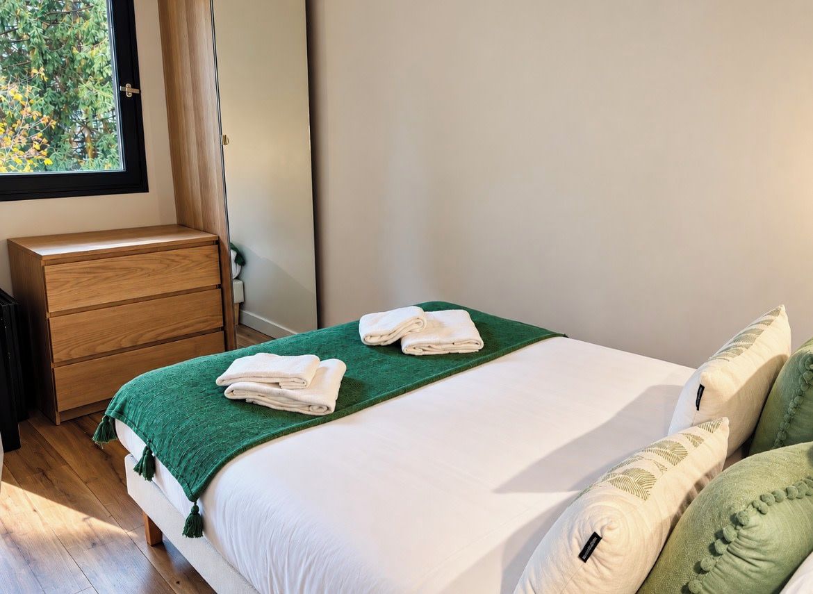 Un letto rifatto con lenzuola bianche e una coperta verde, un comodino in legno e una finestra vista alberi.