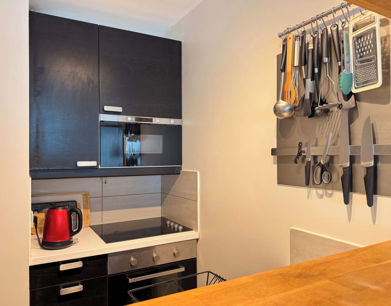 Una cocina compacta con un armario negro, una campana extractora de acero inoxidable, un hervidor rojo y un soporte para utensilios con varias herramientas de cocina.