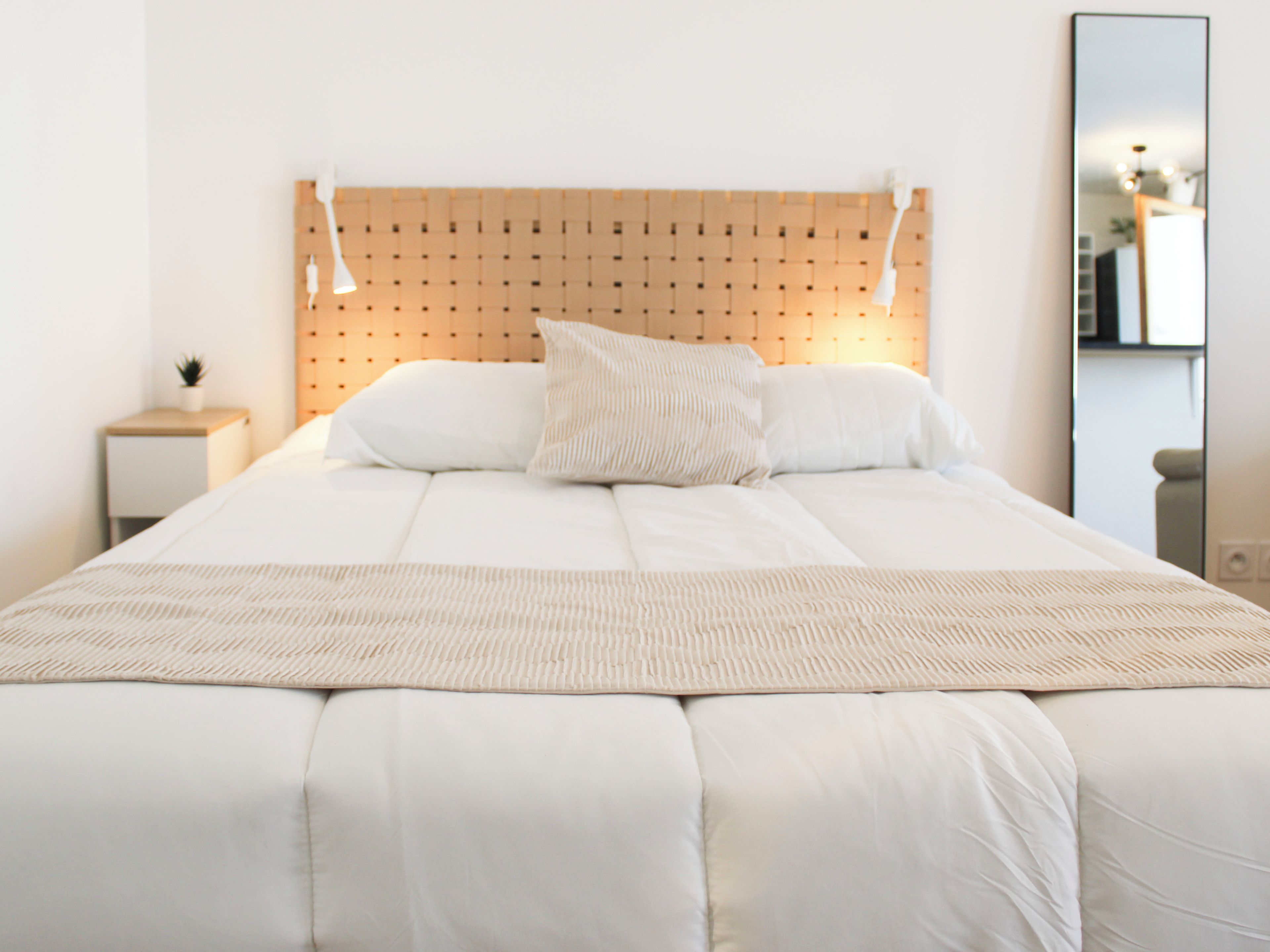 A neatly made bed with a headboard, a bedside table with a lamp, and a mirror on the wall.