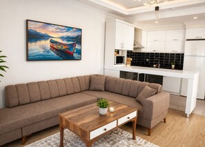 A modern living room with a sofa, a coffee table, and a kitchen area with white cabinets and black backsplash. - Kemer Suite Residence (Antalya)