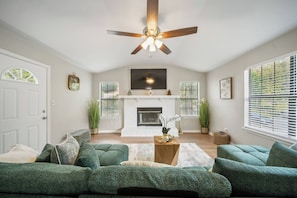 A living room with a fireplace, green sofas, a ceiling fan, and a large window with blinds. - 3BR Near FSU/FAM | Golf Course (Tallahassee)