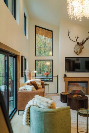 A modern living room with a fireplace, a mounted deer head, and a large window overlooking a forest.