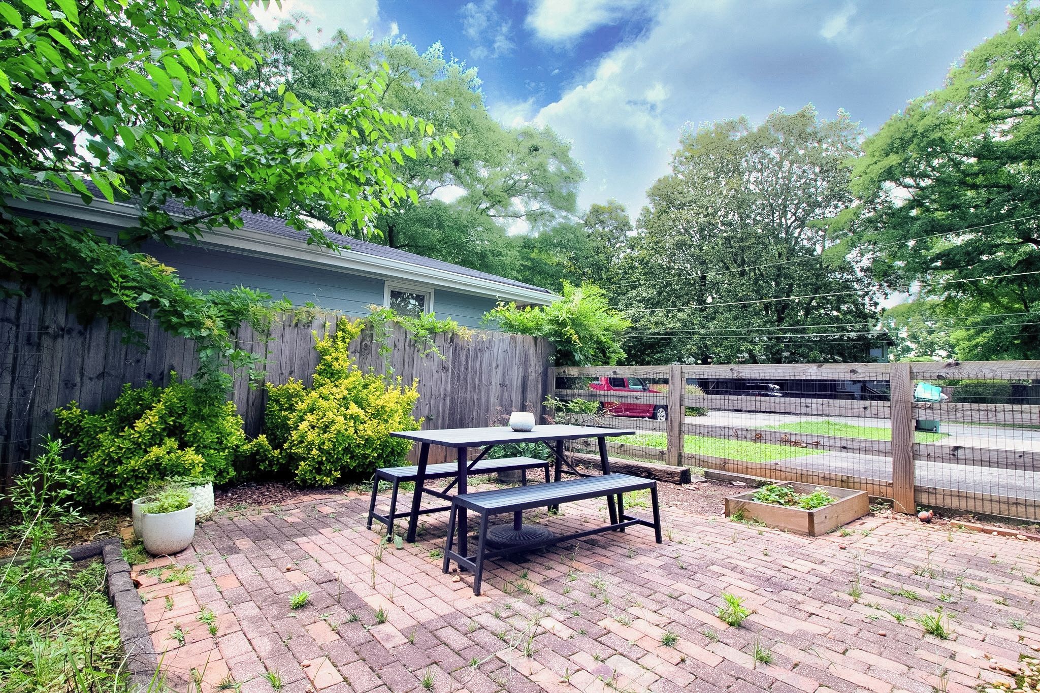Une terrasse à l’arrière de la maison, avec une table et des bancs, entourée d’une clôture en bois et d’une végétation luxuriante.