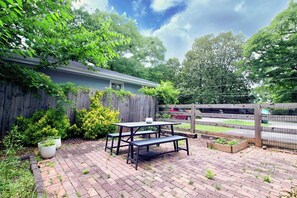 Une terrasse à l’arrière de la maison, avec une table et des bancs, entourée d’une clôture en bois et d’une végétation luxuriante.