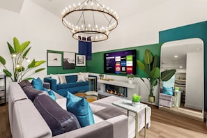 A modern living room with a fireplace, a mounted TV displaying streaming services, and a large mirror reflecting the room. - TWO full houses with a private pool, hot tub, gym, sauna, massage chair, cold plunge, movie projector, astroturf, axe throwing, corn hole and other lawn games! Unwind in this relaxing Paradise Oasis and let your worries drift away! (Austin)