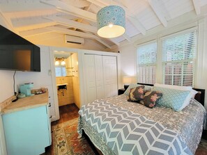 Un dormitorio con cama, mesita de noche, televisor y baño con lavamanos y ventilador de techo.