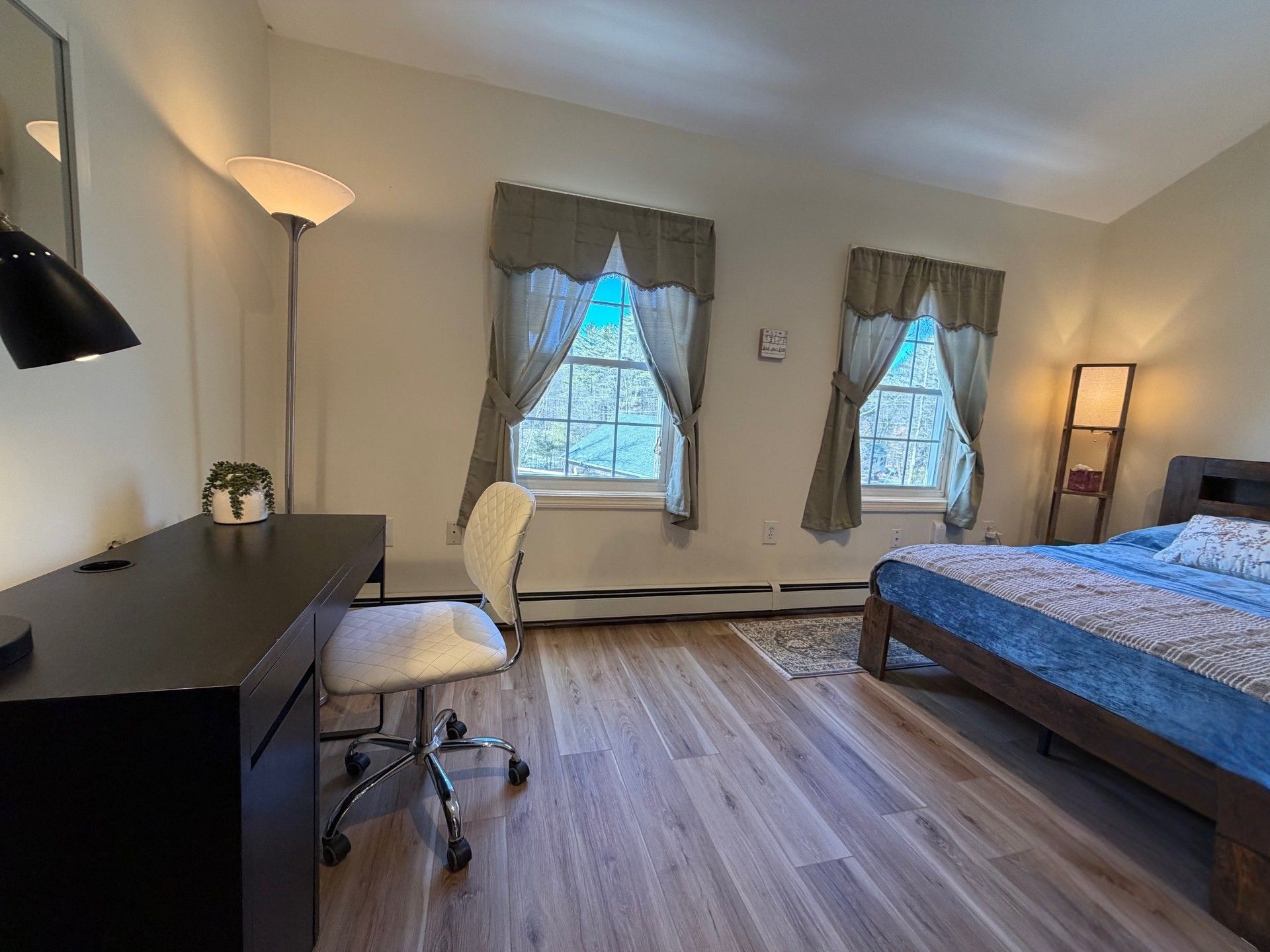 A bedroom with a bed, a desk, a chair, two windows with curtains, a lamp, and a small plant.