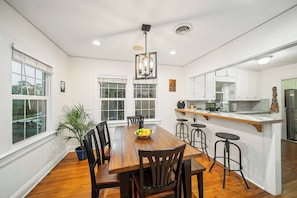 A dining area with a wooden table, black chairs, and a fruit bowl. There are windows with blinds, a ceiling light, and a kitchen area with white cabinets and a stainless steel refrigerator. - 3BR Movie Room | Walk to Dining (Tallahassee)