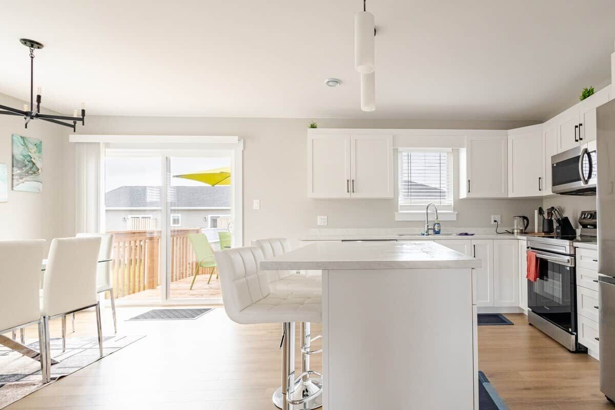 Una cocina moderna con gabinetes blancos, una isla central, electrodomésticos de acero inoxidable y un ventanal corredero que da a un espacio exterior.