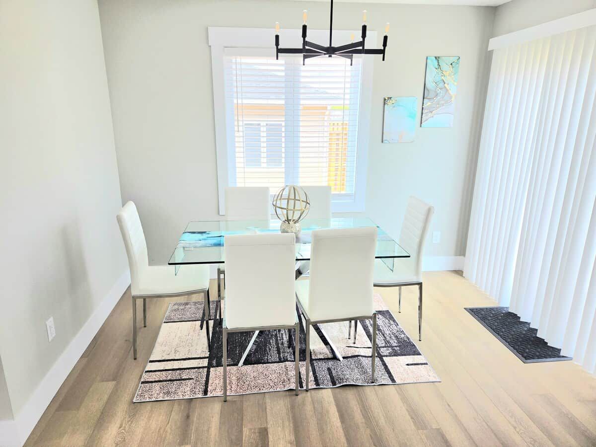 Un coin repas avec une table en verre, des chaises blanches, un tapis noir et blanc, un lustre et une fenêtre avec des stores.