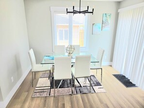 Un coin repas avec une table en verre, des chaises blanches, un tapis noir et blanc, un lustre et une fenêtre avec des stores.