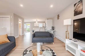 A modern living room with a sofa, a coffee table, and a TV. There is a kitchen in the background.
