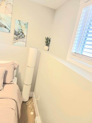 A bedroom with a bed, a window with blinds, a small plant, and abstract art on the wall.
