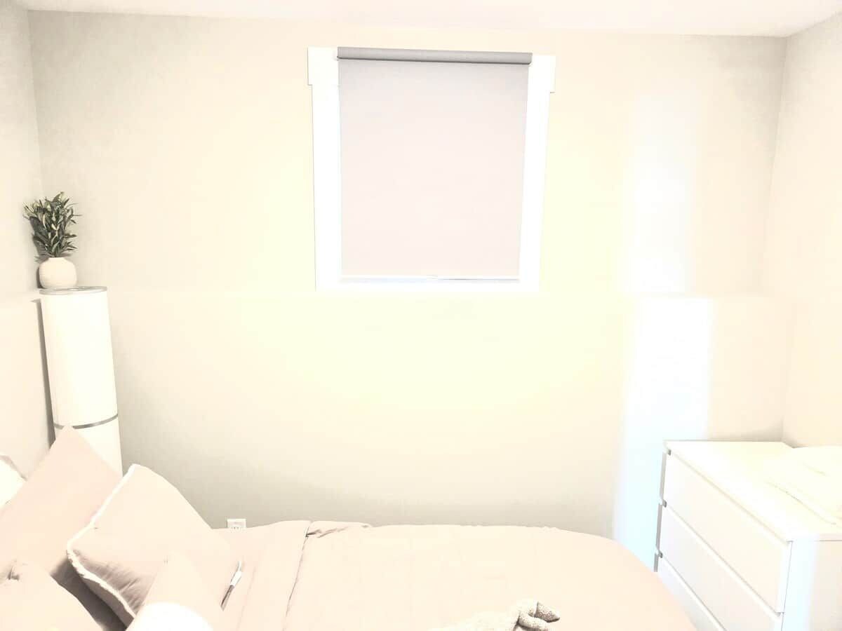 Een slaapkamer met een bed, nachtkastje, lamp, raam met zonwering en een kleine plant.