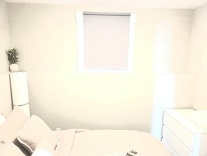 A bedroom with a bed, a bedside table, a lamp, a window with a blind, and a small plant.