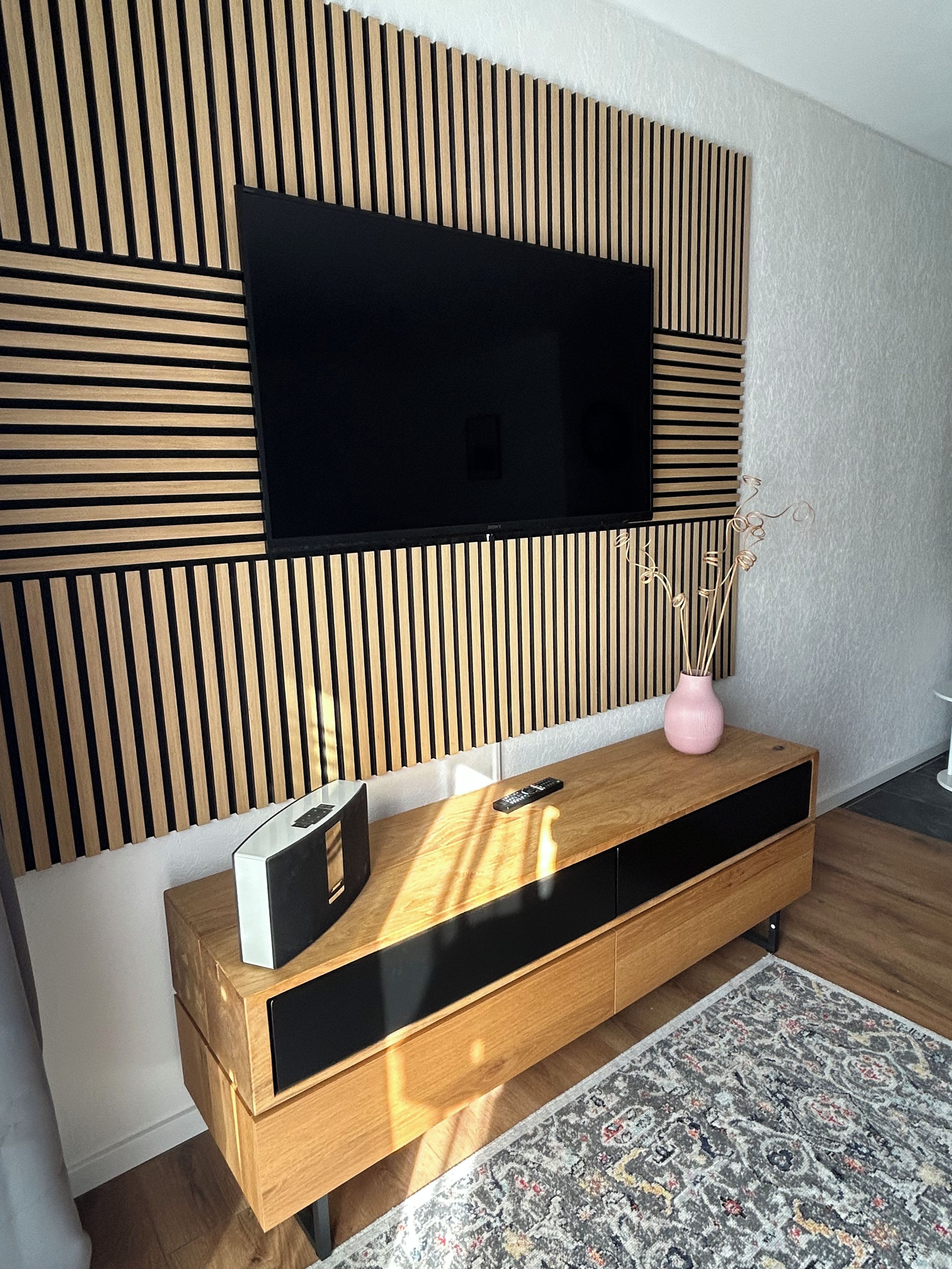 A modern living room with a flat-screen TV mounted on a wooden slat wall, a wooden console table with a remote control, and a patterned rug on the floor.