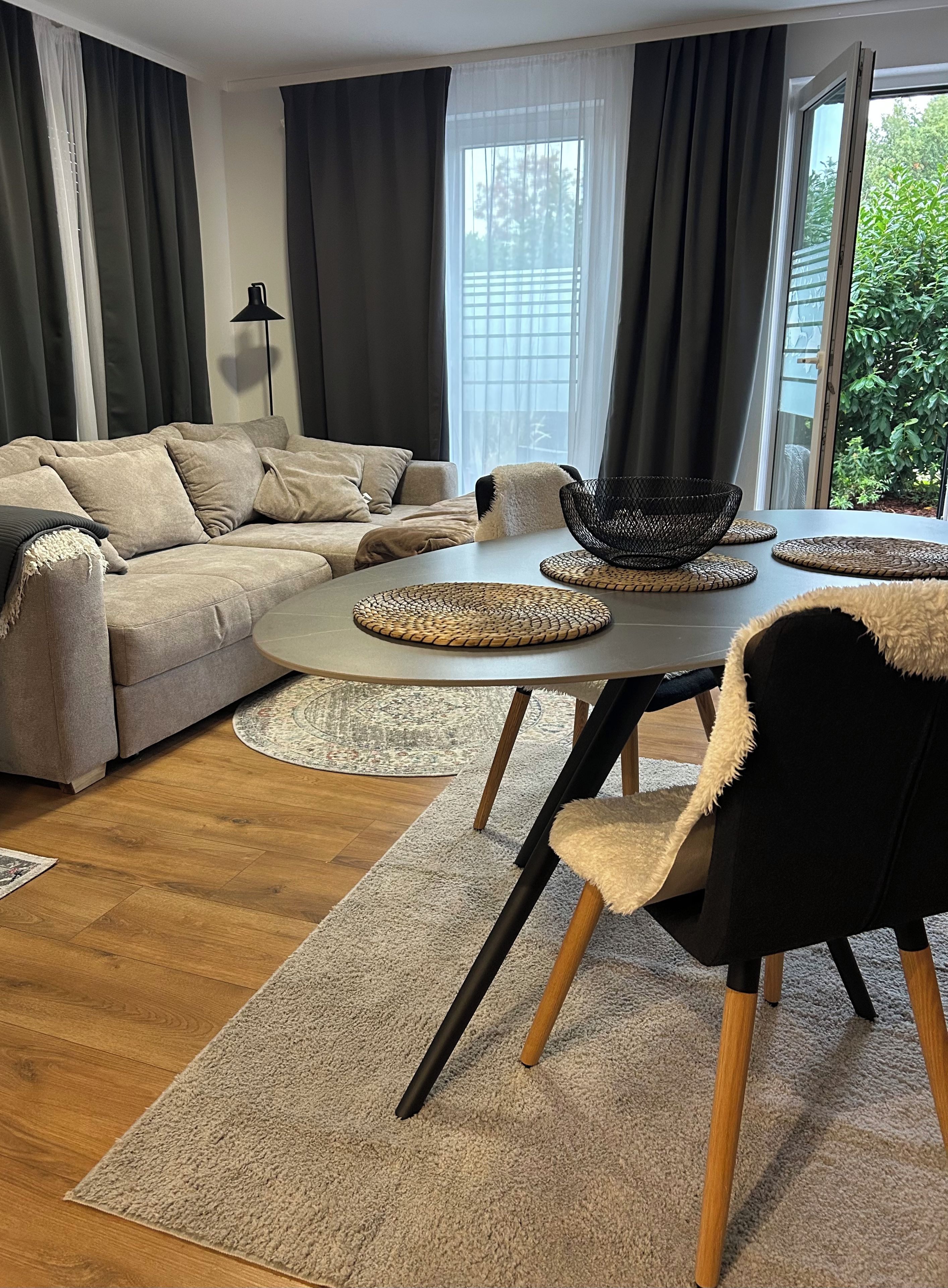 Una sala de estar moderna con un sofá modular, una mesa de comedor redonda con individuales tejidos, y vistas a la vegetación exterior.