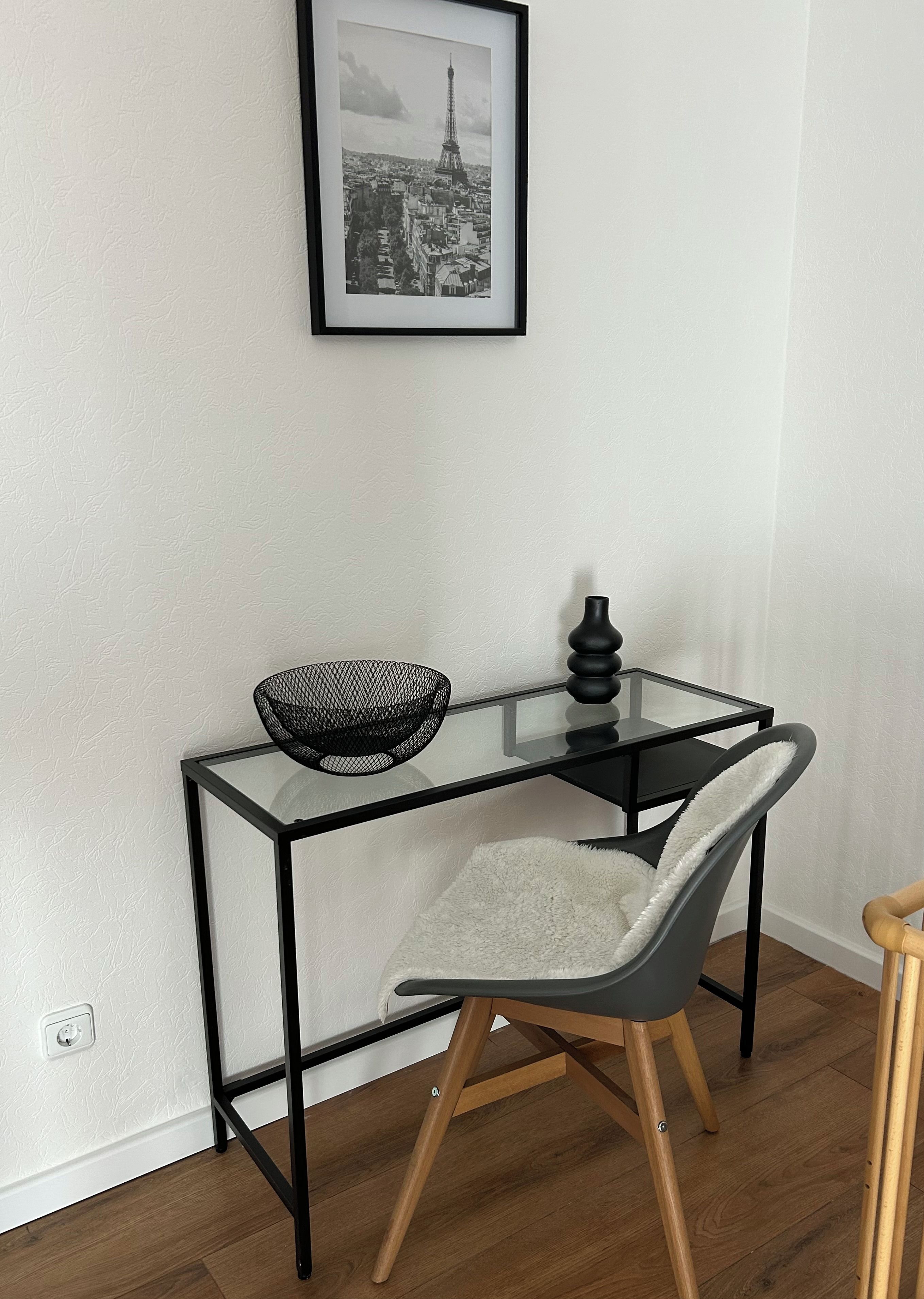 A modern room with a glass-top desk, a chair, a black vase, and a framed picture of the Eiffel Tower.