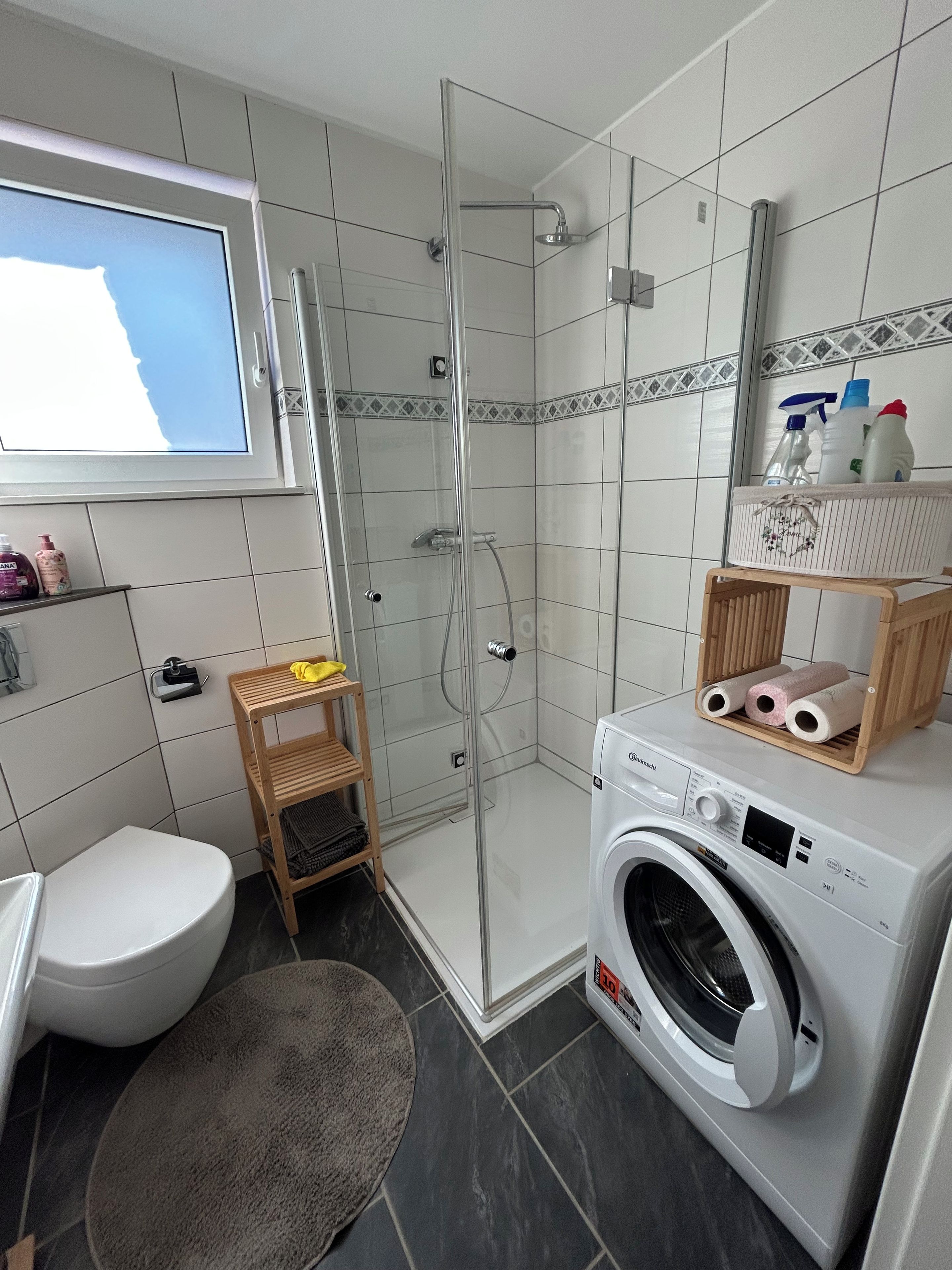 Een moderne badkamer met een douchecabine, toilet, wasmachine en een plank met toiletartikelen.