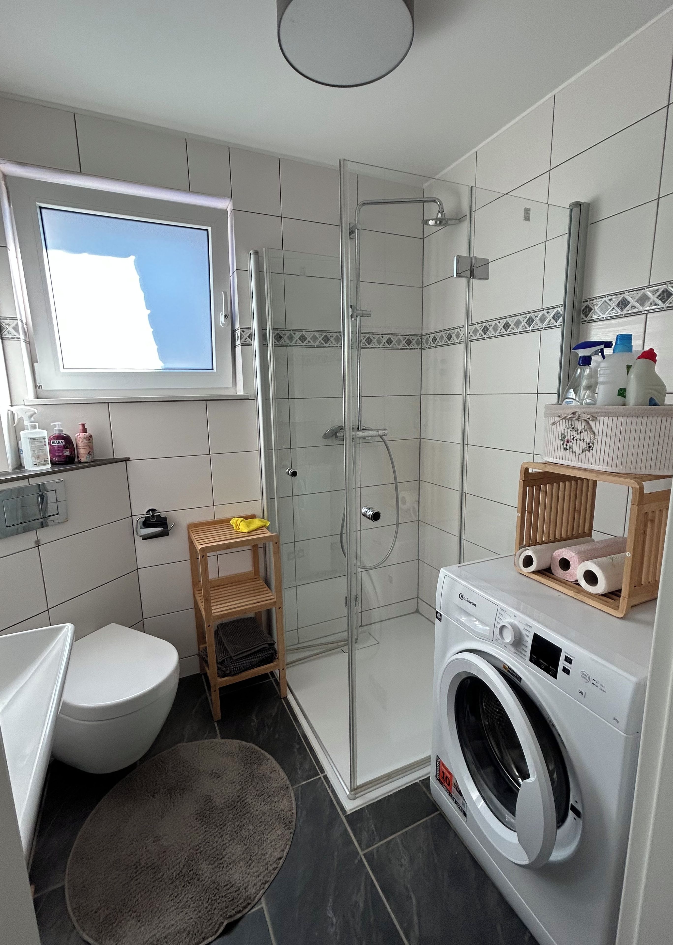 Ein modernes Badezimmer mit einer gläsernen Duschkabine, Toilette, Waschmaschine und einer Holzauslage für Pflegeartikel.