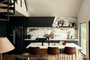 A modern kitchen with a marble backsplash, black cabinets, and a dining area with leather chairs. - The Coler Treehouse - Steps to MTB Trails, 1 Mi to Downtown Bentonville (Bentonville)