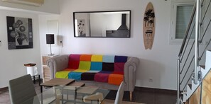 Ein Wohnzimmer mit einem farbenfrohen Sofa, einem Glastisch, einem Sessel, einem Kamin, einem Wandspiegel und einer Surfbret-Dekoration.