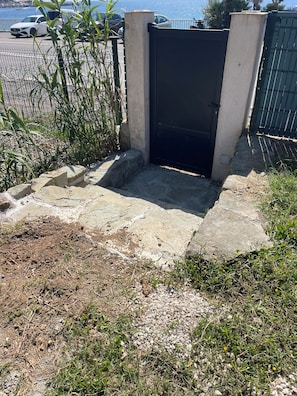 A gated entrance with a concrete pathway leading to a dark door, surrounded by vegetation and a parking area in the background. - An exceptional villa with a pool, right by the sea, combining tranquility and elegance. (Sainte-Maxime)