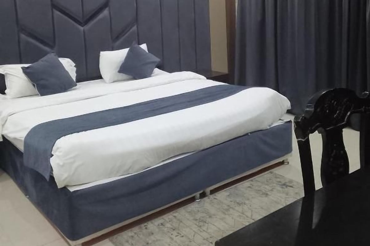 A modern bedroom with a large bed, a headboard with geometric patterns, a dark curtain, and a small table.