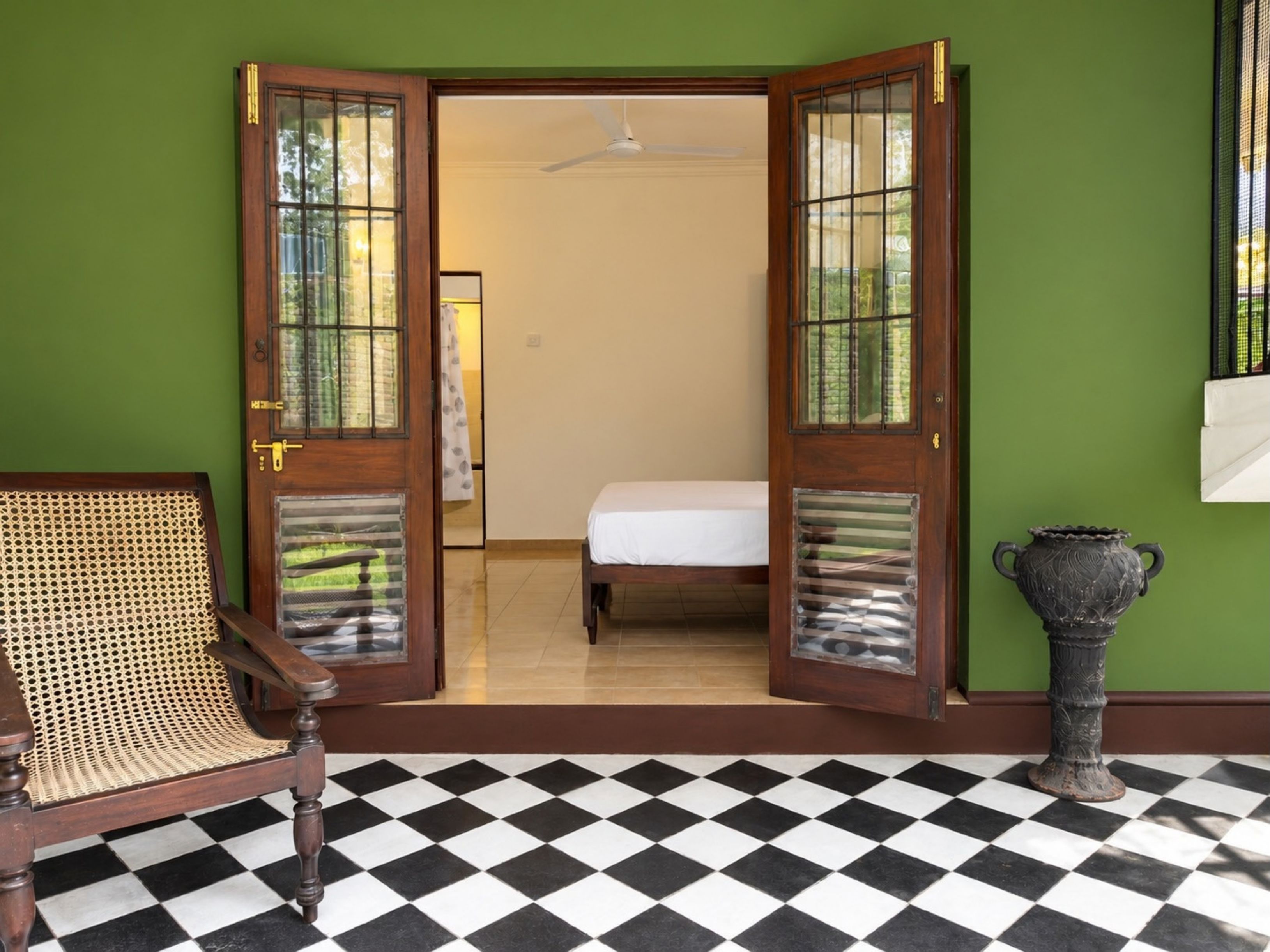 A room with a checkered floor, a bed, a rattan chair, and a decorative urn.