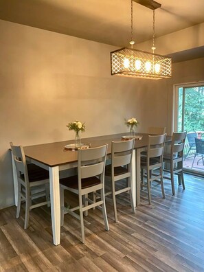 Un coin repas comprenant une table rectangulaire et six chaises, un luminaire suspendu et une vue sur l’extérieur grâce à une grande fenêtre.