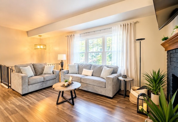 A living room with two sofas, a coffee table, a fireplace, and a large window with curtains.