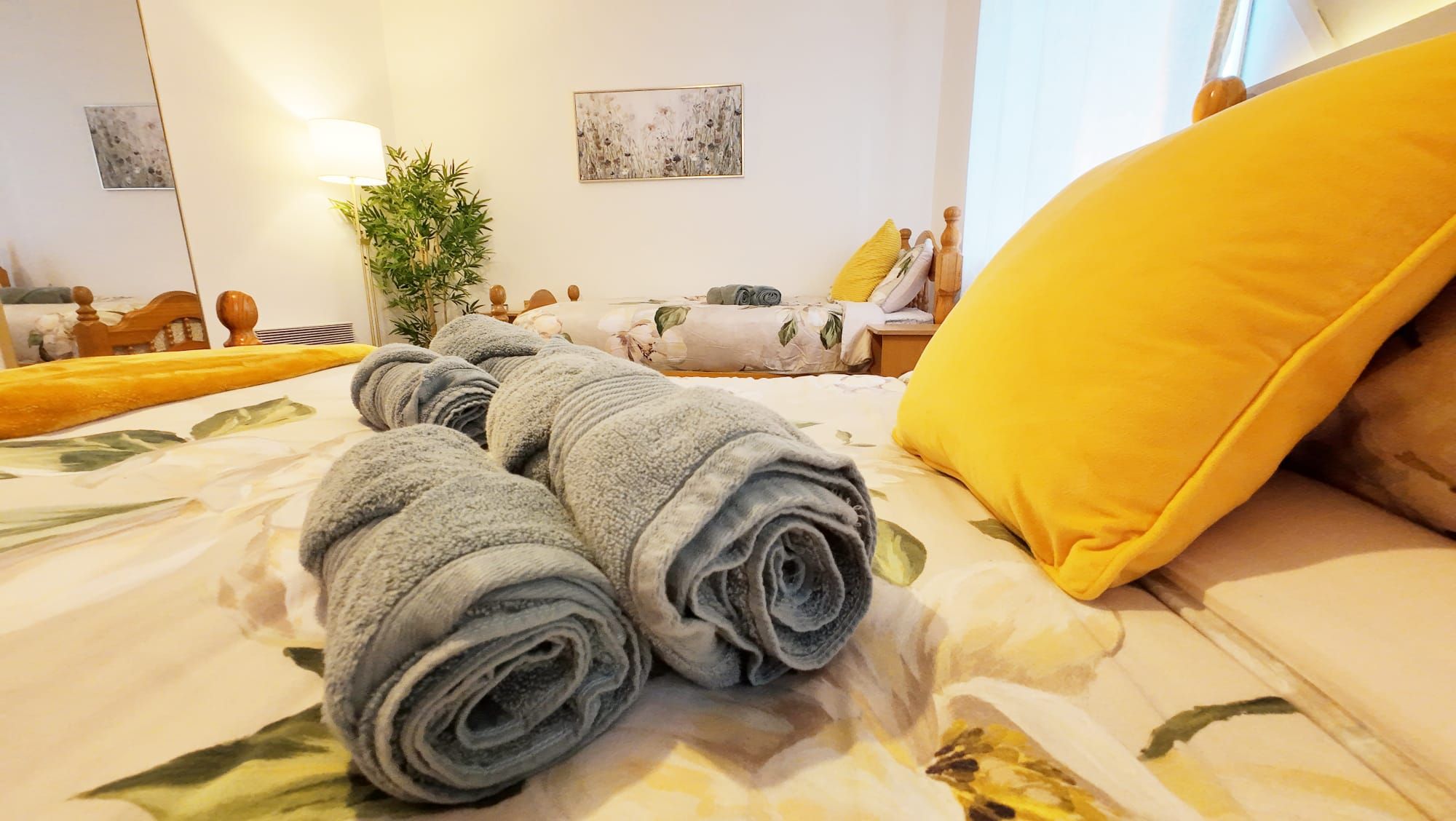 A neatly made bed with rolled towels, a yellow pillow, and a plant in a room with a mirror and artwork on the wall.