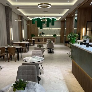 Un moderno vestíbulo de hotel con mesas redondas, asientos cómodos, una recepción y elementos decorativos colgantes verdes.