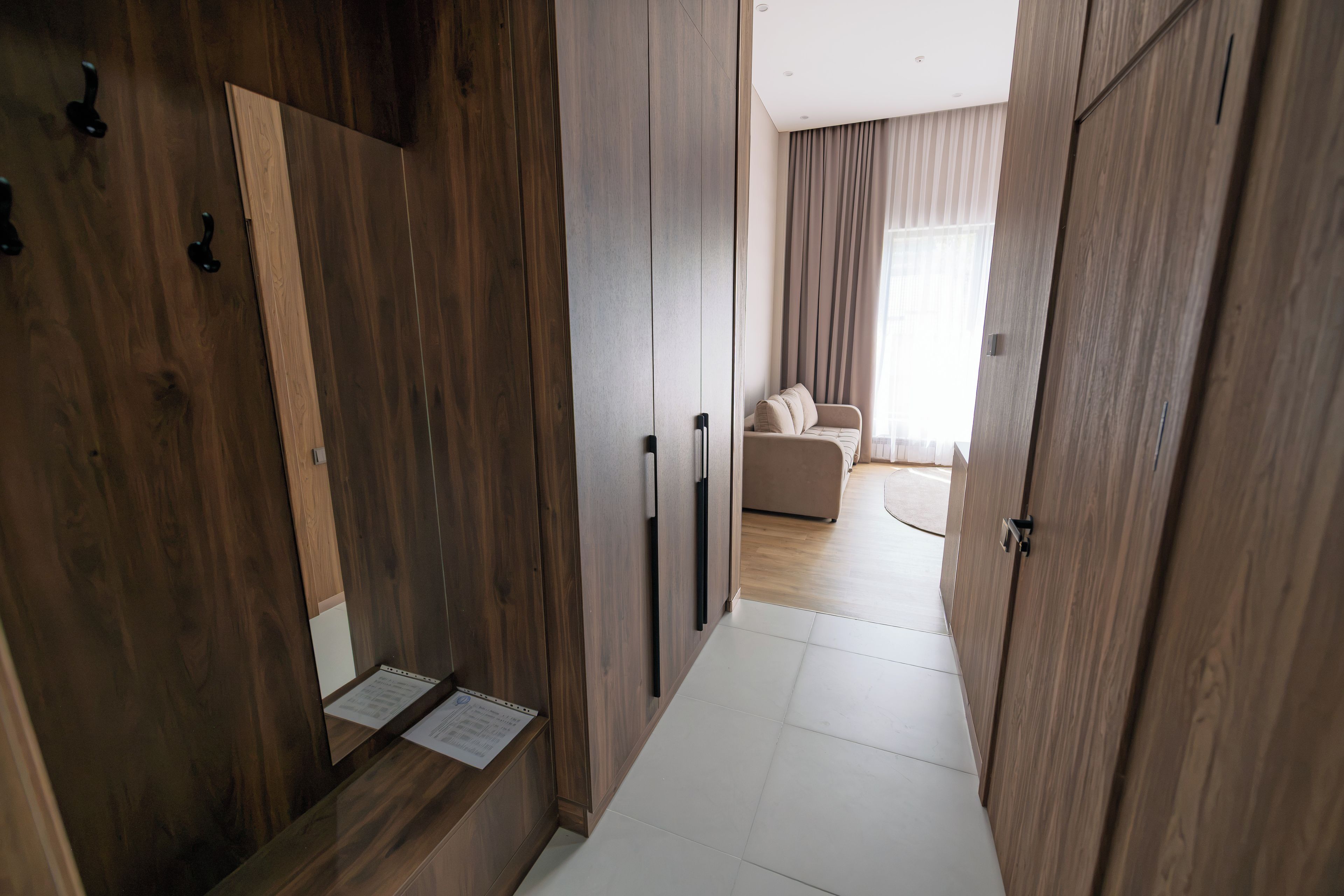 A hallway with wooden doors, a white tiled floor, and a view into a living area with a sofa and curtains.