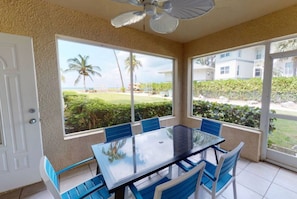 A room with a glass table and blue chairs, large windows with a view of a lawn and palm trees, and a ceiling fan. - Family friendly seven mile beach condo.Cayman gem . (George Town)