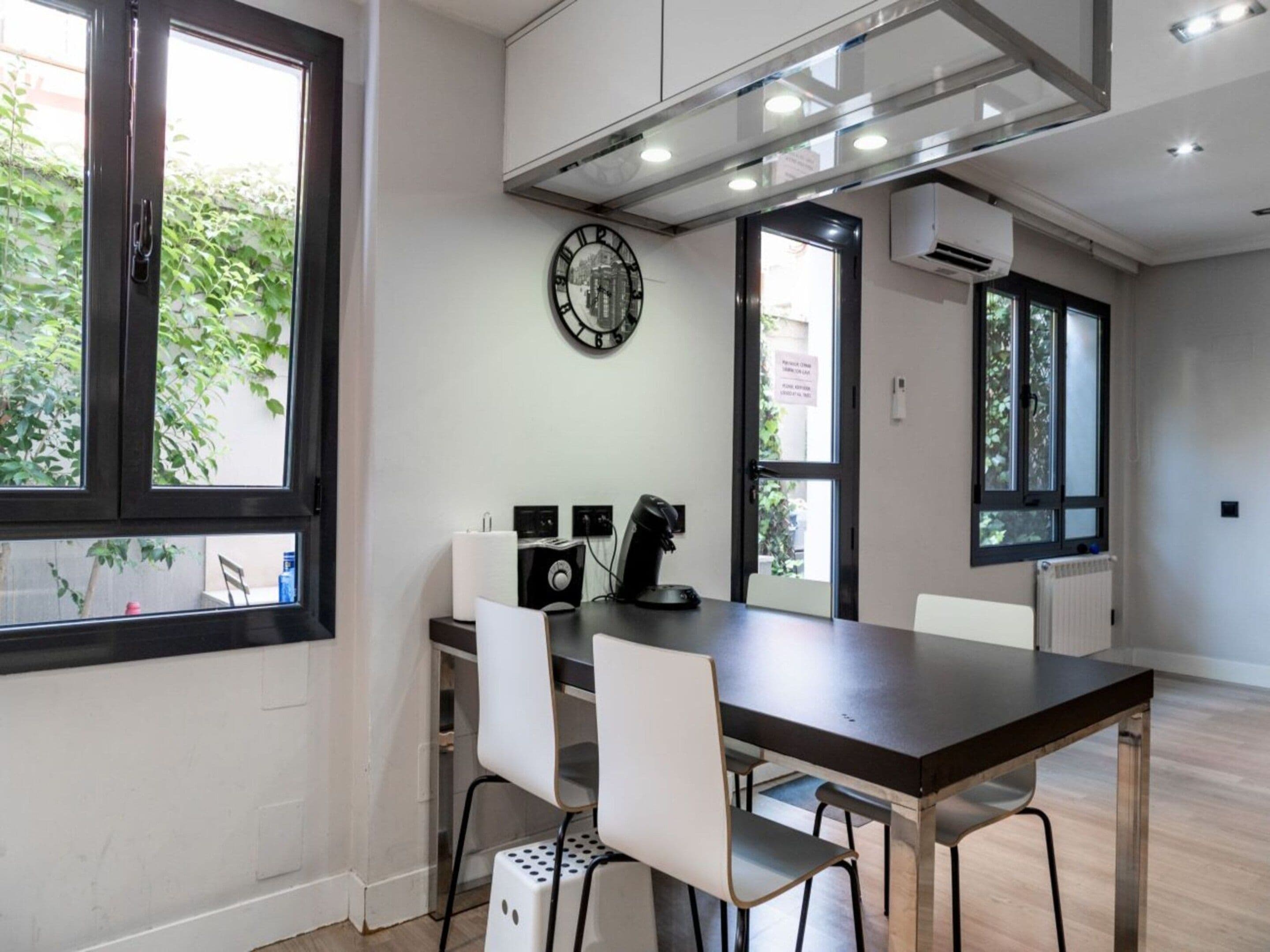 A modern kitchen with a dining table, chairs, a clock, and a view of greenery outside.