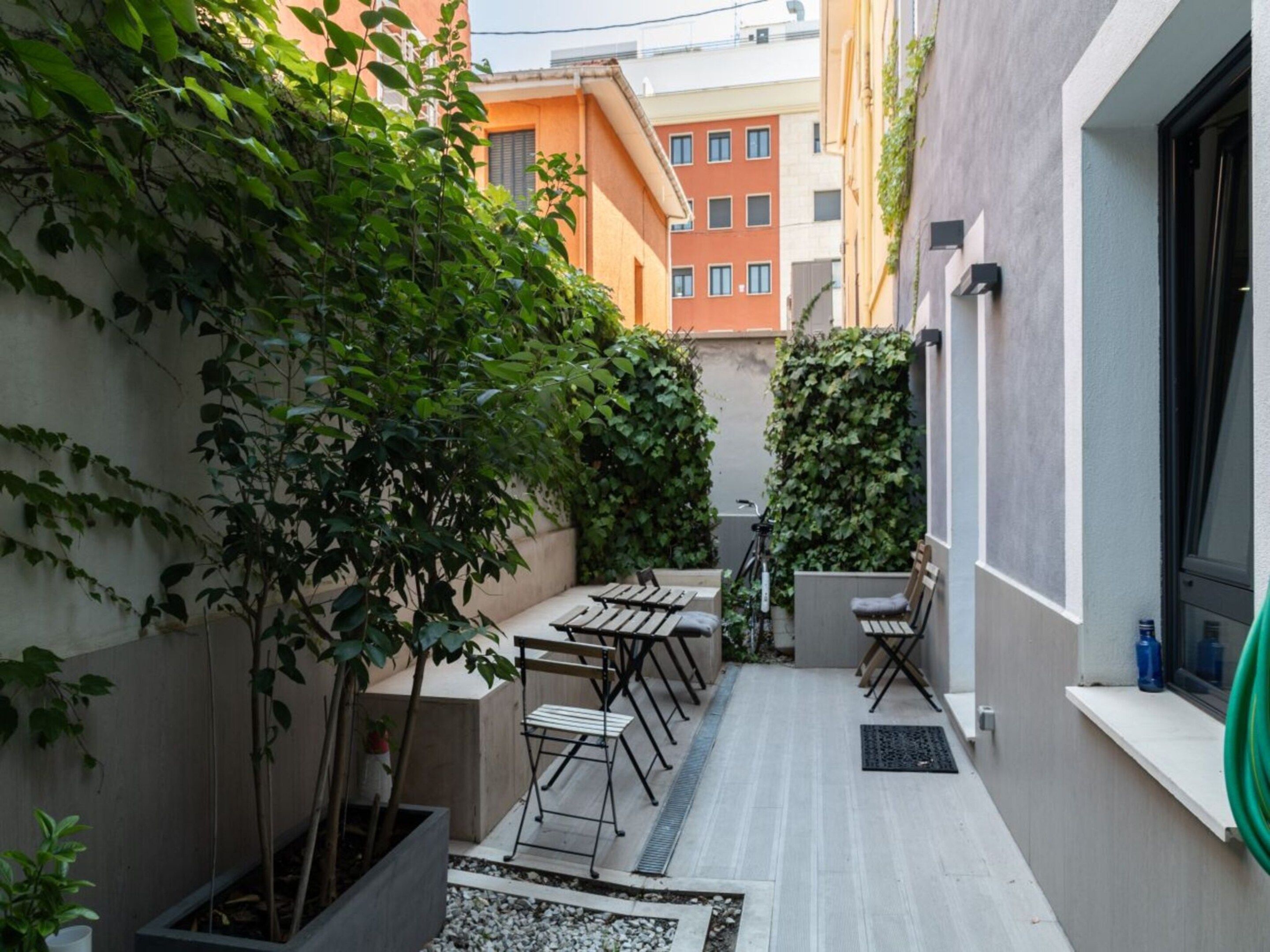 A narrow outdoor patio with a seating area, a bicycle, and a hose on the right.