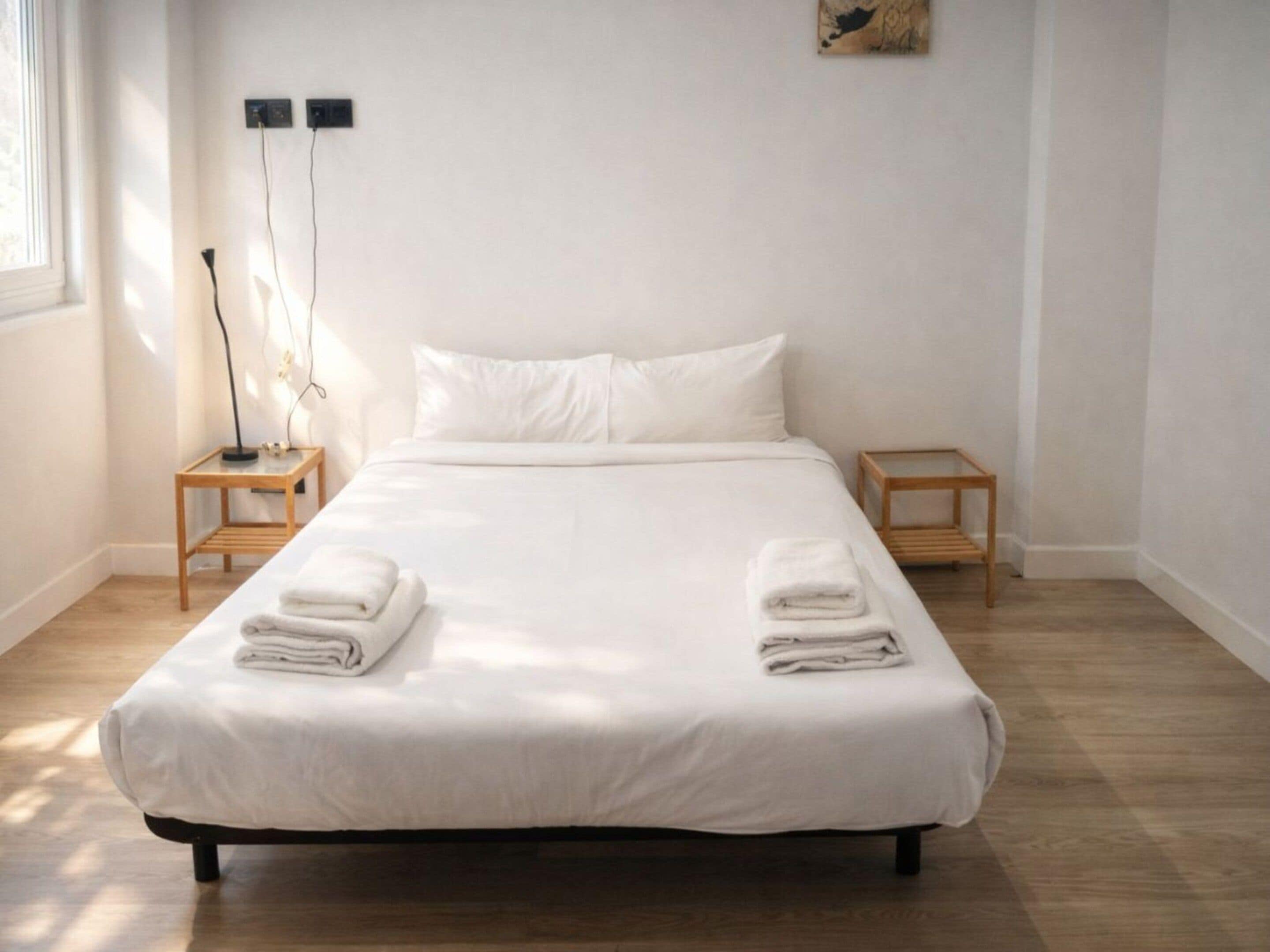 A neatly made bed with white linens and two folded towels on the bed. There are two bedside tables with lamps, and a wall-mounted power outlet above the bed.