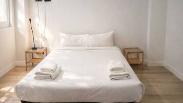 A neatly made bed with white linens and two folded towels on the bed. There are two bedside tables with lamps, and a wall-mounted power outlet above the bed.