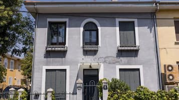 A two-story building with a symmetrical facade, featuring three windows with shutters, a central entrance with a door, and a small balcony on the upper floor.