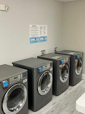 Un espace buanderie comprenant quatre machines à laver à chargement frontal, une affiche fixée au mur avec des instructions pour le lavage du linge, et un comptoir blanc.