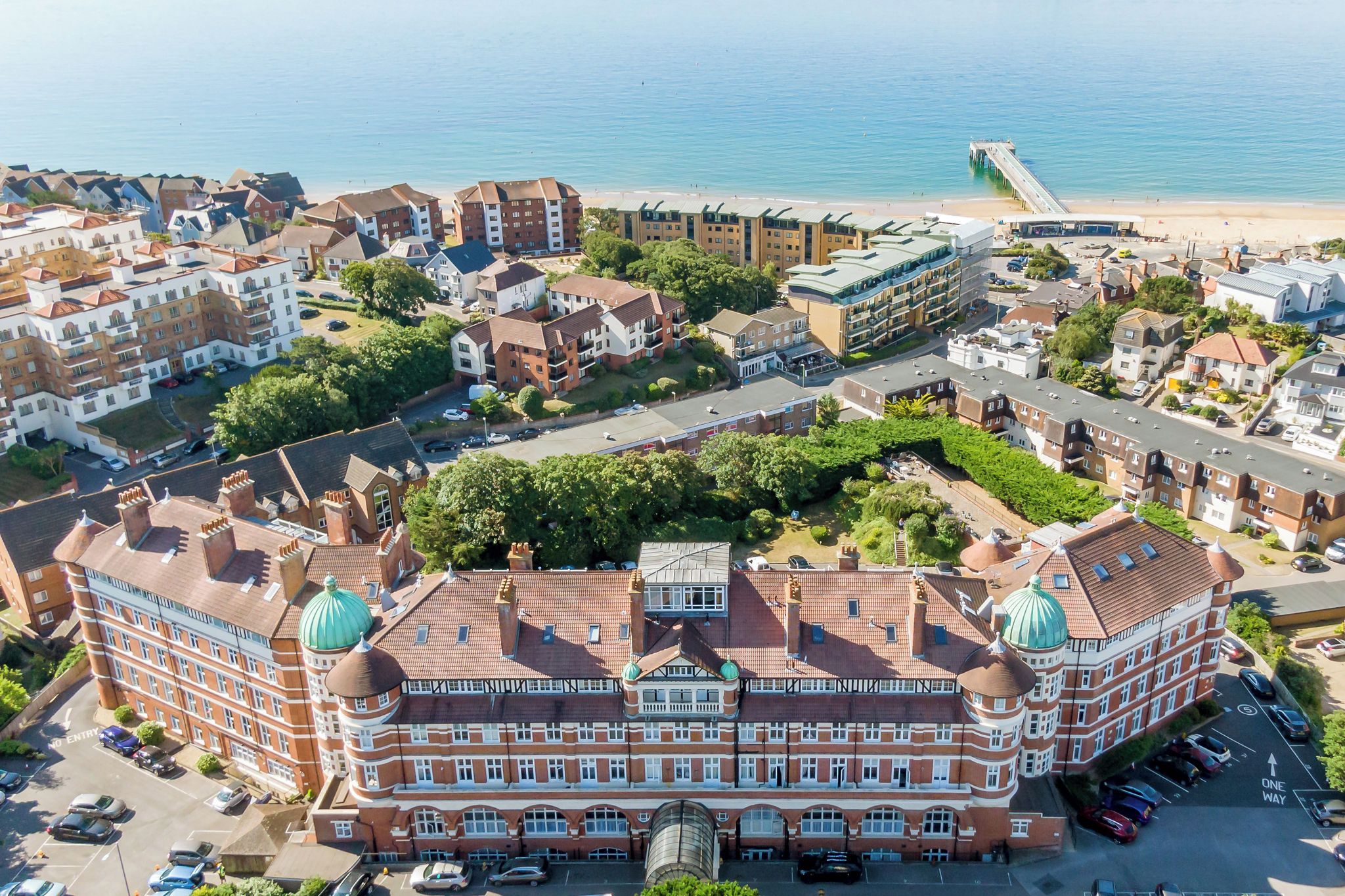 Um grande edifício histórico com vários andares e telhados verdes distintos em forma de cúpula, cercado por um estacionamento e uma praia.