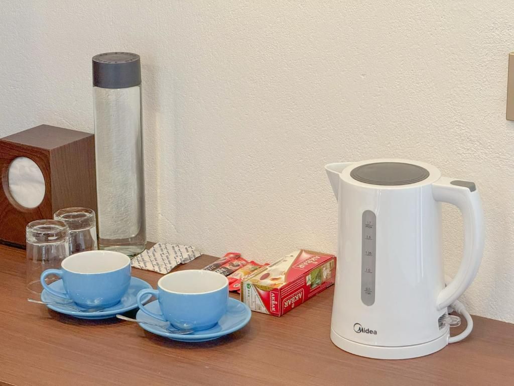 Una mesa con un hervidor eléctrico blanco, dos tazas azules con platillos, un vaso de agua, un termo y una caja de bolsitas de té.