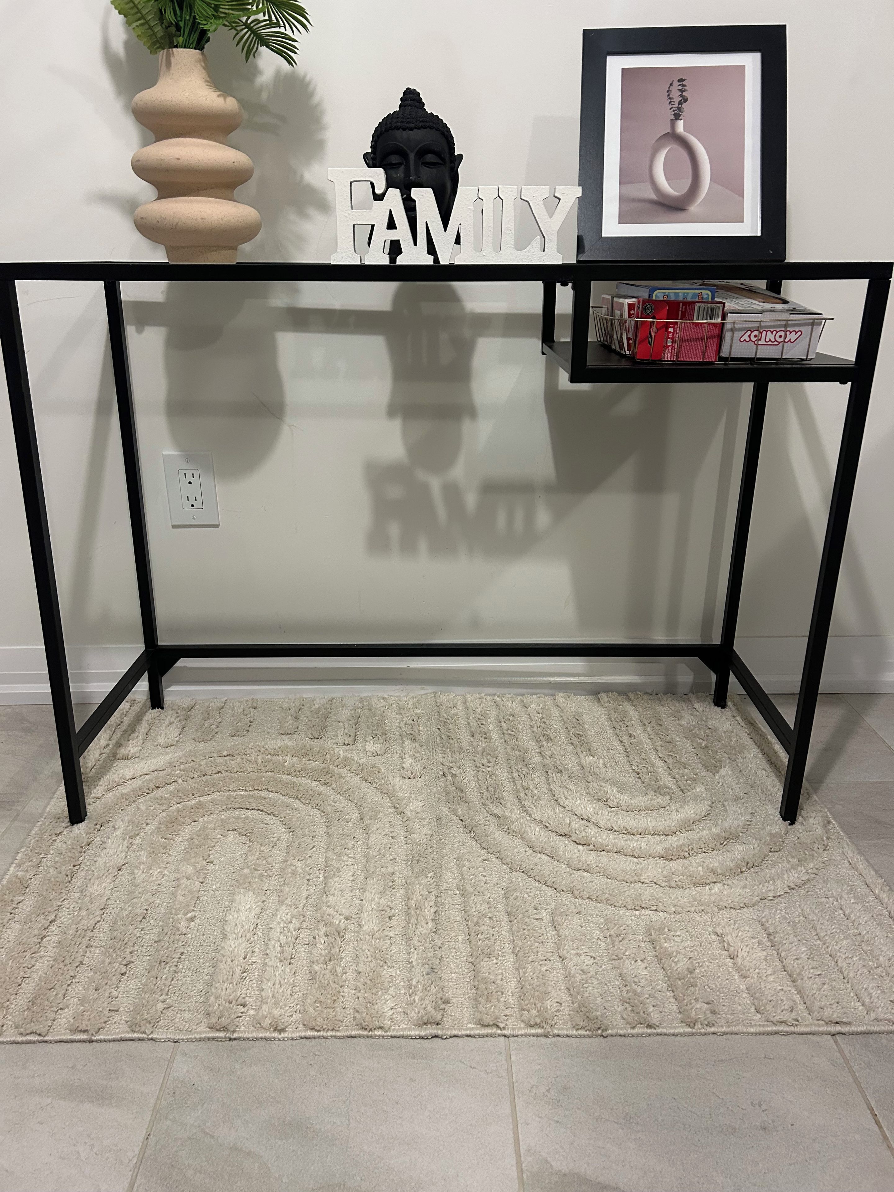 Une pièce avec un bureau à plateau de verre, un vase beige orné de feuillage vert, un tableau encadré et le mot « FAMILLE » écrit sur le mur.
