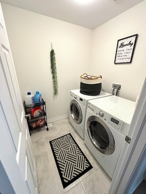A laundry room with two front-loading washing machines, a small sink, a rack with laundry supplies, and a decorative wall hanging.