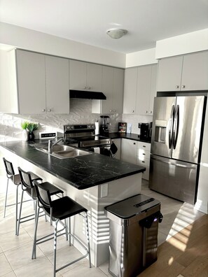 A modern kitchen with a central island, stainless steel appliances, and bar stools. - The Comfy Couch | Modern 3BR HOME+ Private Jacuzzi (Haldimand)